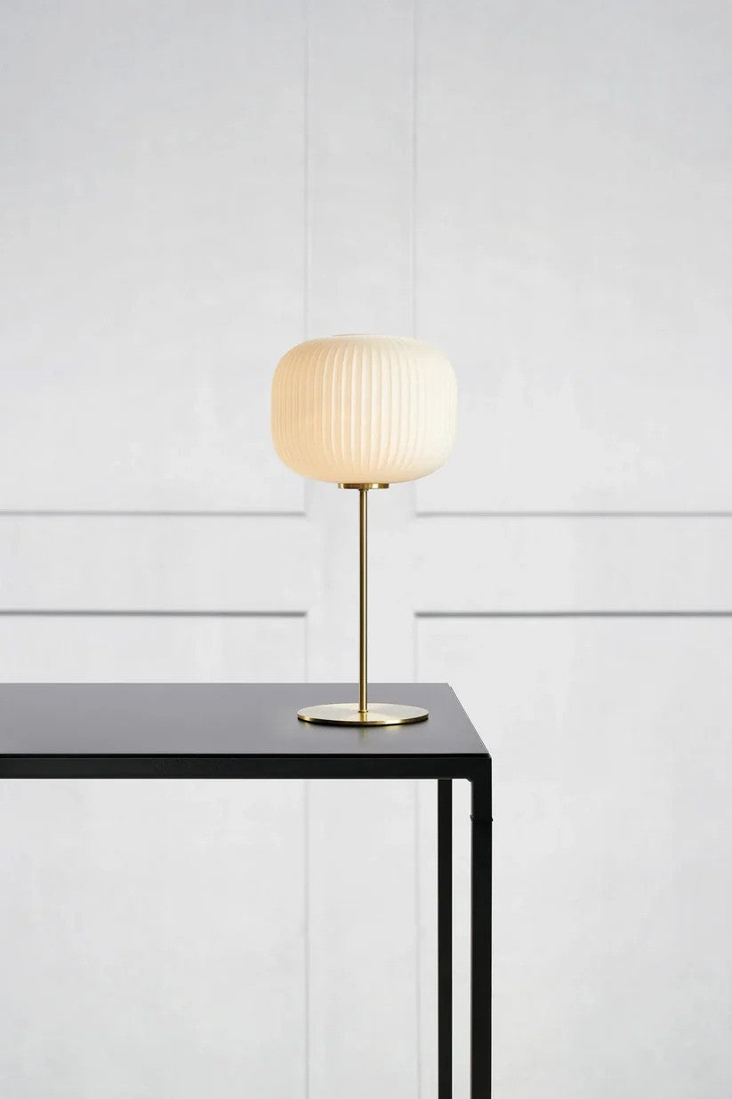 Sober Bordlampe i Messing/Hvid placeret på et bord med væg bagved for den flotte kontrast af den elegante bordlampe