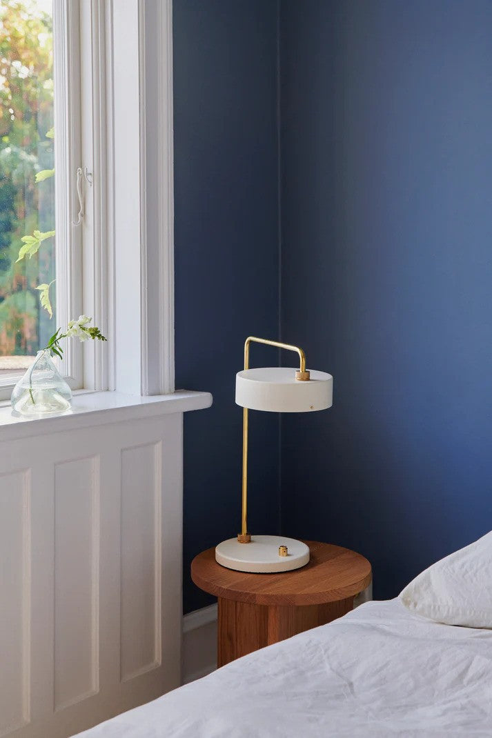Den elegante Petite Machine Bordlampe Hvid placeret på et bord ved sengekanten med dens varme belysning og nemme betjening