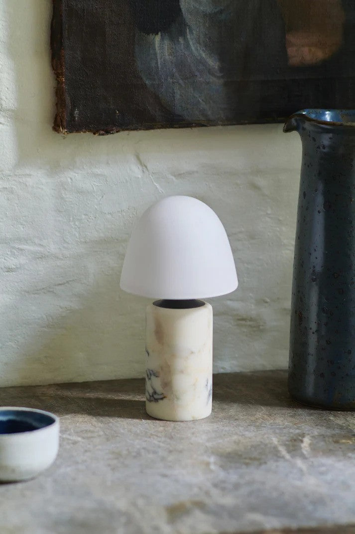 Den æstetiske Fuse Portable Bordlampe placeret på hylden foran en hvid væg for en flot kontrast og dens elegante design