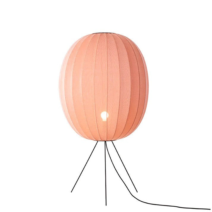 Den æstetiske Knit-Wit Ø65 High Oval Floor Medium i Coral placeret foran en hvid væg med en E27 pære iført for den behagelige belysning